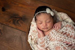 A three week old newborn baby girl sleeping in a little wooden bowl. She is wearing a cream colored bow headband and swaddled with a decorative wrap. Mothers and new parents can get the help they need for postpartum depression therapy in New Jersey. Brave Minds Psychological Services in Scotch Plains, NJ will help you get postpartum depression treatment.