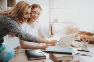Mother looks over college daughter's shoulder as she points to something on her laptop. They are at a cluttered dining room table and smiling. Brave Minds Psychological Services offers online therapy for college students in new jersey, anxiety scotch plains, nj, and more! Contact us today!