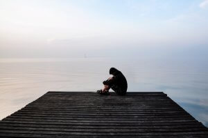 Frustrated depression man in the hood sitting on wooden bridge near the beach on sunset. Could be suffering from COVID stress of unemployed sadness depressed and human problems with PTSD and Trauma. Trauma therapy and PTSD Treatment in Scotch Plains, NJ are available at Brave Minds Psychological Services Counseling Clinic and in online therapy in New Jersey