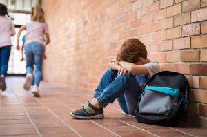 Little boy sitting against a brick wall with his backpack and arms on his legs with head in his arms. School anxiety makes his scary thoughts worse. He needs help from a child psychologist in Scotch Plains, NJ or in online therapy for children in New Jersey.