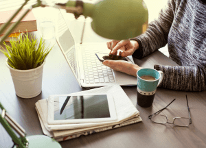 A light skin toned person sits at a table working on their laptop. There is a tablet, newspaper, a cup of coffee, a pair of glasses, and a small plant on the table as well.