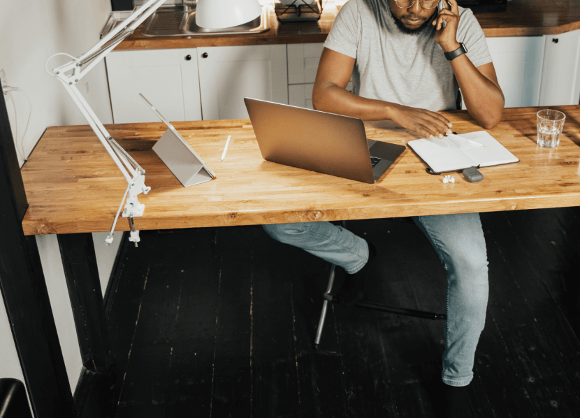 A medium-dark skin toned man sitting at a desk in his kitchen workin on his laptop. He is talking on his cell phone.