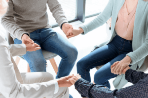 A group counseling session. Four members are sitting close together holding hands. You see their legs and arms only.