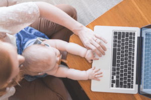 A new mom holds her baby on her lap. The baby is playing with the mother's laptop.