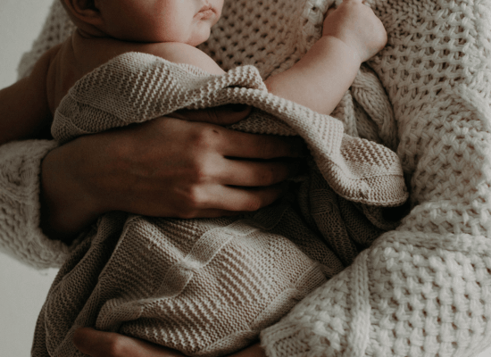 A mother holds her newborn baby close to her. The baby is wrapped in a cream colored blanket.