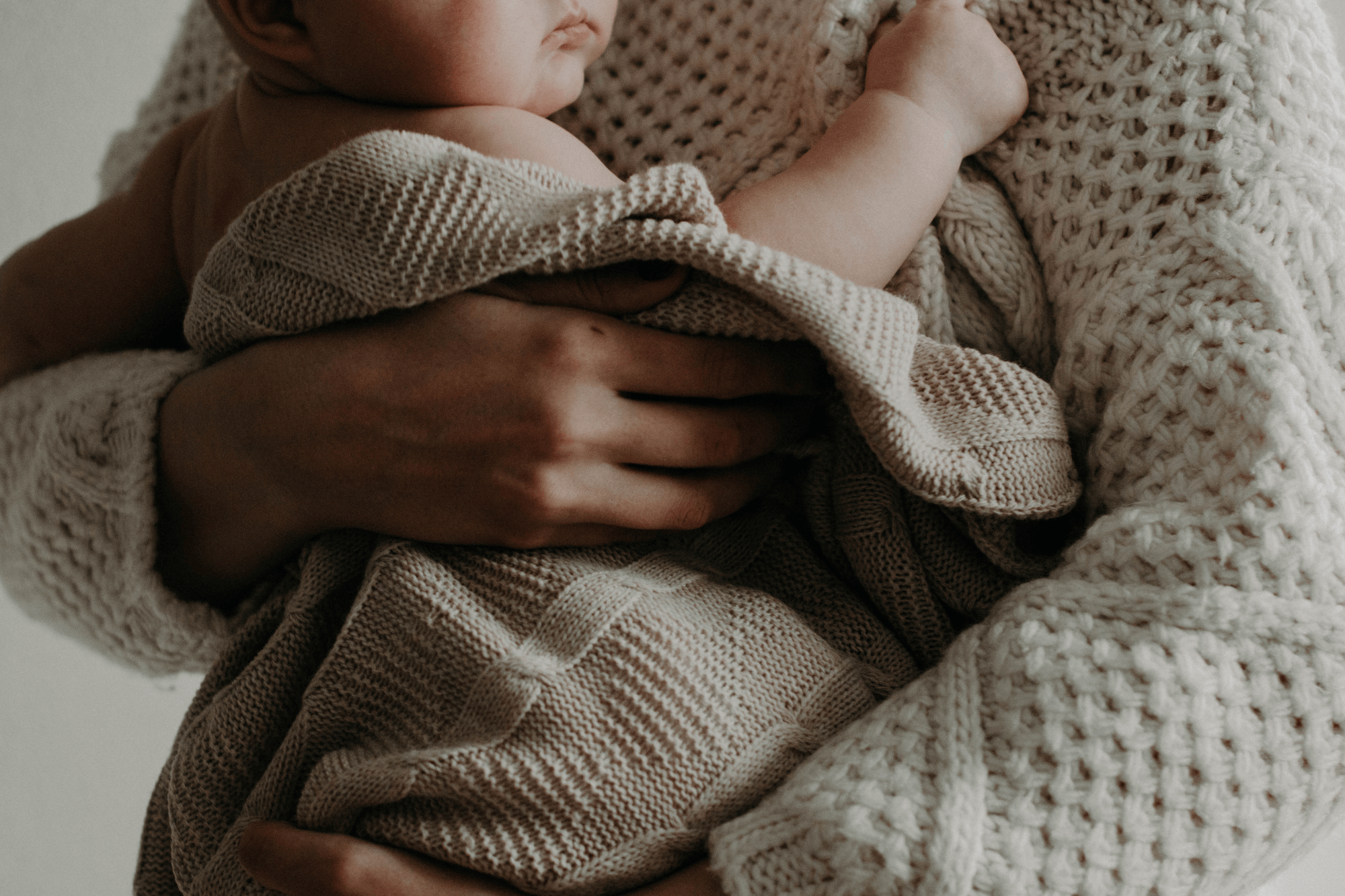 A mother holds her newborn baby close to her. The baby is wrapped in a cream colored blanket.