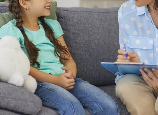 A close-up of a woman smiling with a clipboard while sitting next to a child. This could represent the support offered by a child psychologist during child therapy for anxiety. Learn more about the benefits of child counseling in Scotch Plains, NJ today.