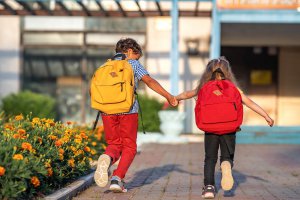 Two kids with backpacks run toward a school while holding hands. Learn how a child psychologist can offer support for you and your child. Search “child therapy online in New Jersey” to learn more about child therapy for anxiety today.