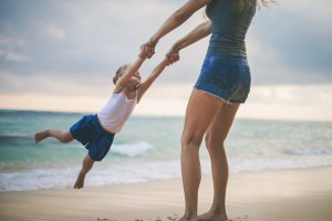 A parent swings their child by the arms on the shore. This could represent the stronger bonds cultivated through child therapy for anxiety. Contact a child psychologist to learn more about child therapy online in New Jersey and other services. 