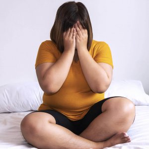 woman covering her face wearing a yellow shirt sitting on a bed
