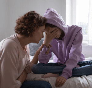 A light skin toned mother is speaking her to her teenage daughter. The daughter is sitting on her bed with her face in her hands. Their foreheads are touching.