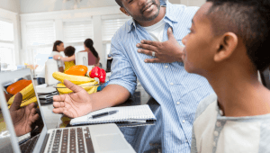 A medium dark skin toned man has a conversation with his son in their kitchen. His son is sitting in front of a laptop with a notebook and pen next to him.