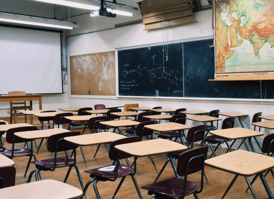 An empty classroom full of desks. There is writing on the chalk board and a map hanging on the wall.