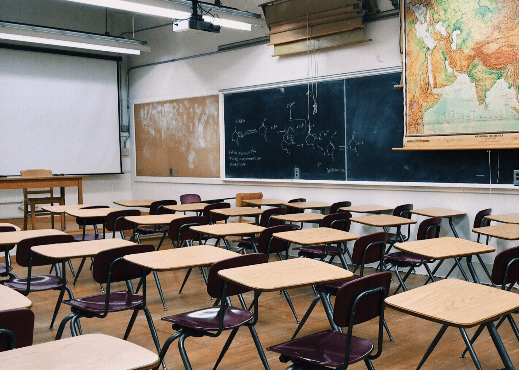 An empty classroom full of desks. There is writing on the chalk board and a map hanging on the wall.