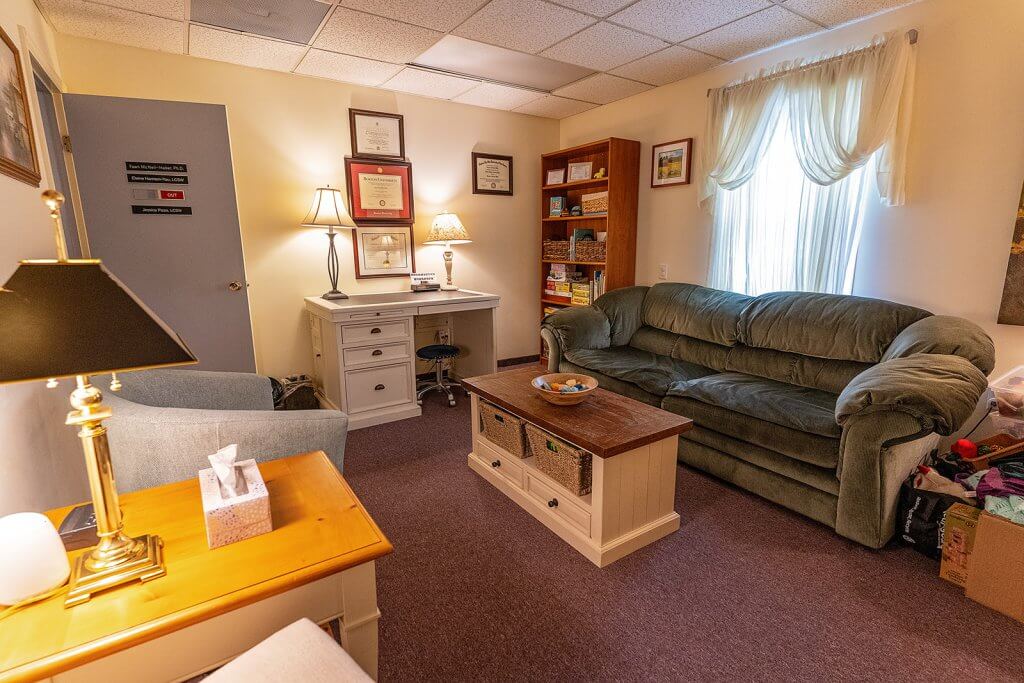 A therapist's office or an office where a Mental Health Administrative Assistant would work. There is a green couch, a brown coffee table, a gray chair, and a white desk. Behind the couch there is a window with white curtains hanging.