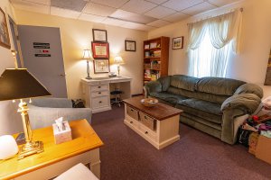 A therapist's office or an office where a Mental Health Administrative Assistant would work. There is a green couch, a brown coffee table, a gray chair, and a white desk. Behind the couch there is a window with white curtains hanging.