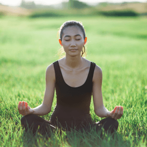 A light skinned young woman meditating in a field. She is sitting in the grass with her eyes closed.