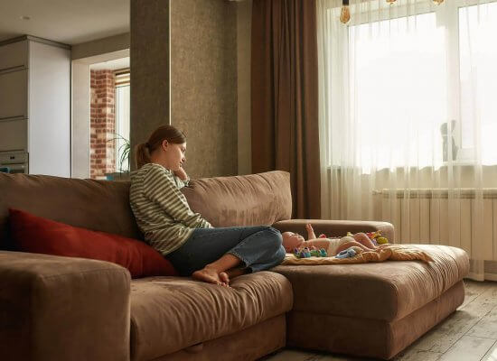 Sad mother sitting on a couch watching her infant. Call today to speak with a trauma therapist in Scotch Plains, NJ.