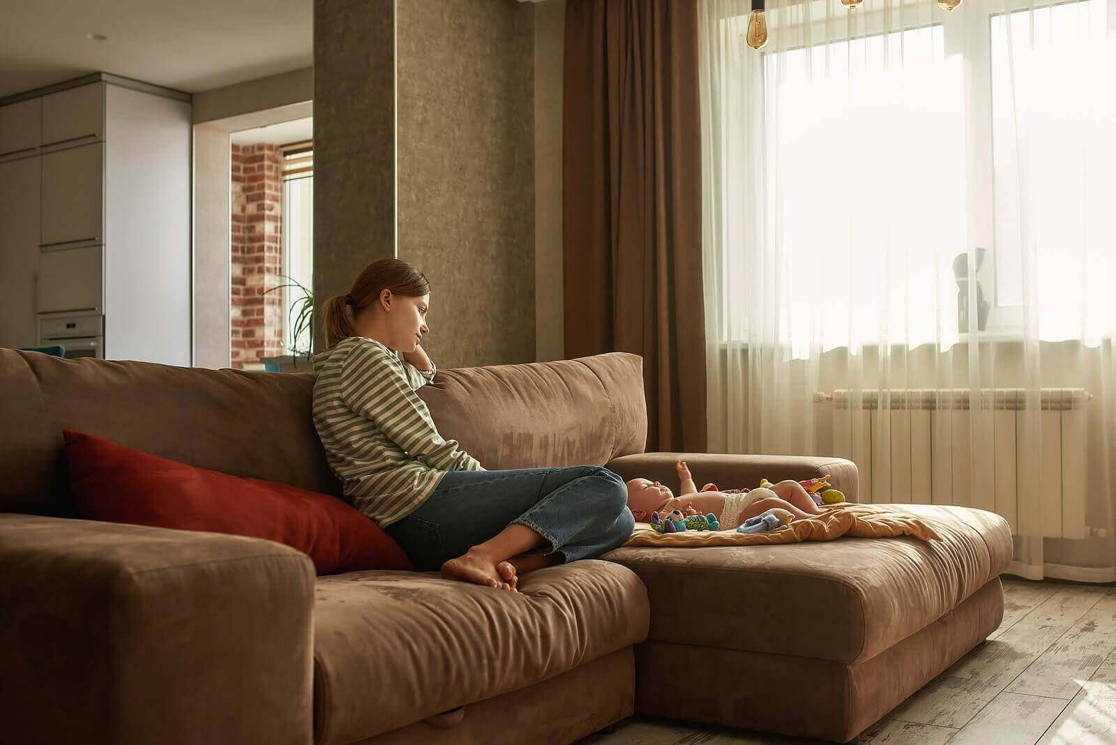 Sad mother sitting on a couch watching her infant. Call today to speak with a trauma therapist in Scotch Plains, NJ.