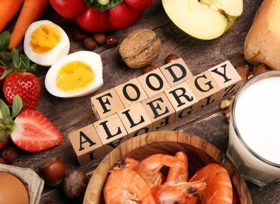 Image of food allergy spelled out in wooden blocks surrounded by food that could cause anaphylactic reaction in New Jersey. Thus showing that just the image of foods can cause food allergy anxiety but a trauma therapist can help with that.