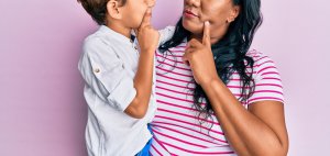 Image of a mother holding her son in front of a pink background with both of them holding a finger to their cheeks. This represents the struggle of knowing that to do next to help your child with selective mutism. For a parent who feels stuck a child psychologist or therapist can work with you and your Childs school.