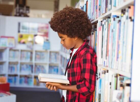 Image of a boy in a red shirt reading a book at a school library. Showing how selective mutism can affect how kids socialize at school. However through child counseling a child psychologist can make a plan for the school that support your kid.