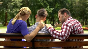 Two parents talk with their child on a park bench. This could represent a difficult conversation that parent coaching in Scotch Plains, NJ can offer support addressing with your child. Search "trauma therapist in Scotch Plains, NJ" to learn more about antiracism in Scotch Plains NJ today.