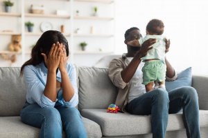 A mother covers her face while her husband holds their child on the couch. Contact a child therapist in Branchburg, NJ to learn more about the benefits of child therapy in Branchburg, NJ, and other support for new parents.