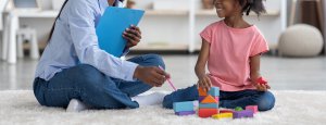 A child playing with blocks smiles while listening toa woman holding a clipbaord. This could represetn the support a child therapist in Branchburg, NJ can offer. Learn more about EMDR for children and other services by searching for child therapy in New Jersey today.