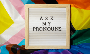 Different flags of the LGBTQIA+ community overlayed with a felt board and writing that reads, "Ask My Pronouns." Understanding the transitions of transgender youth can be difficult. Seek therapy with a transgender youth counselor to help guide the process.