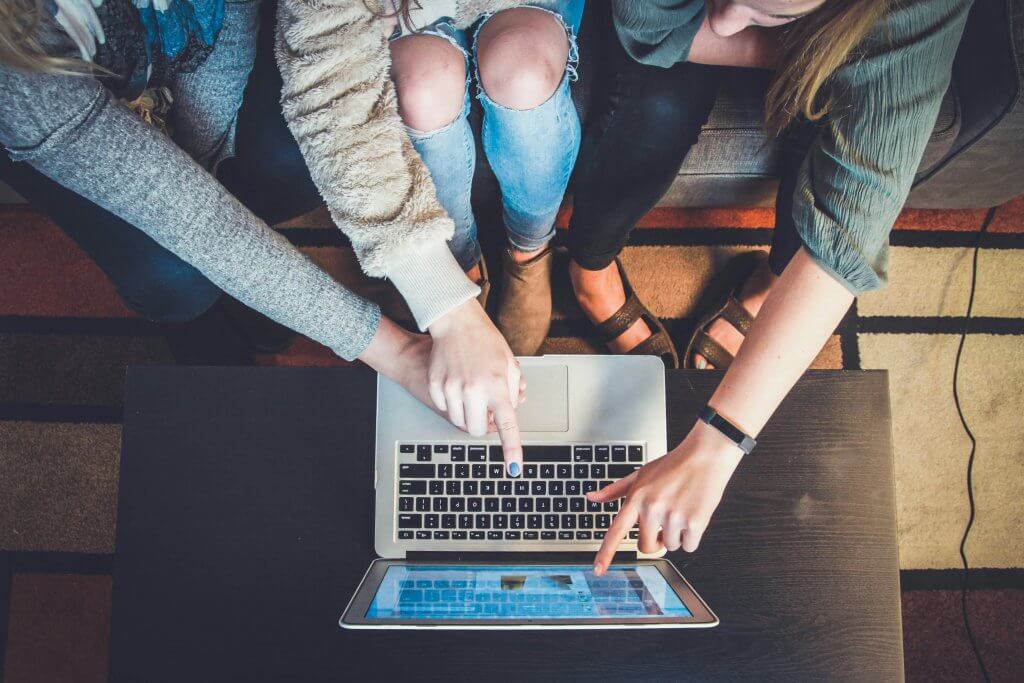Three women using a laptop on a wooden table, talking about how helpful it can be to join a mom support group online in New Jersey. Contact us today to learn more about our Braving Motherhood group for moms.