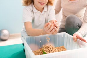 A child smiles while playing with a tub of beads. This could represent the support that child therapy for anxiety can offer for addressing stress. Search for child counseling in Branchburg, NJ and the help a child therapist in Branchburg, NJ can provide.