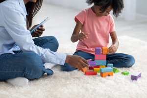 A black woman takes notes on a clipboard while a child plays with blocks. This could symbolize the support child therapy in New Jersey can offer. Learn more about child counseling in Branchburg, NJ and the help child therapy for anxiety can offer. 