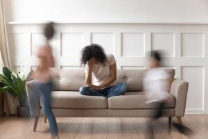 A mother sits on a couch with a stressed expression as her kids run around her. This could symbolize the stress of parenting that a mom support group online in New Jersey can address. Learn more about online group therapy in New Jersey today by searching for a group therapist in Scotch Plains, NJ today. 