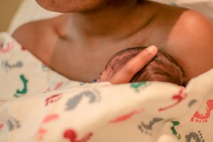 An infant resting against a mom's chest, covered by a blanket with colorful prints, during an online support group meeting.