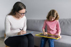 A woman takes notes on a clipboard while a child draws on a chalkboard. This could symbolize the support child therapy in New Jersey can offer. Learn more about child counseling in Branchburg, NJ and the help child therapy for anxiety can offer.