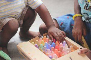 Two children sitting on the floor selecting chalk from a box as part of their at-home anxiety treatment. Look for a play child therapist in New Jersey today!