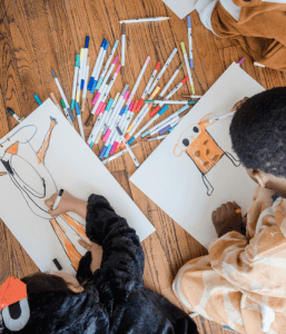 Two children in one-piece pajamas drawing pictures with markers on the floor as part of their social play to help with child anxiety. Look for a child play therapist near you.
