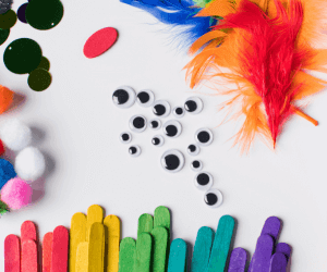 Colorful craft materials for child anxiety treatment including popsicle sticks, googly eyes, pompoms, feathers, and glitter on a white background.