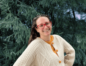 A smiling woman in red glasses and a cream cable-knit sweater stands in front of green pine trees.