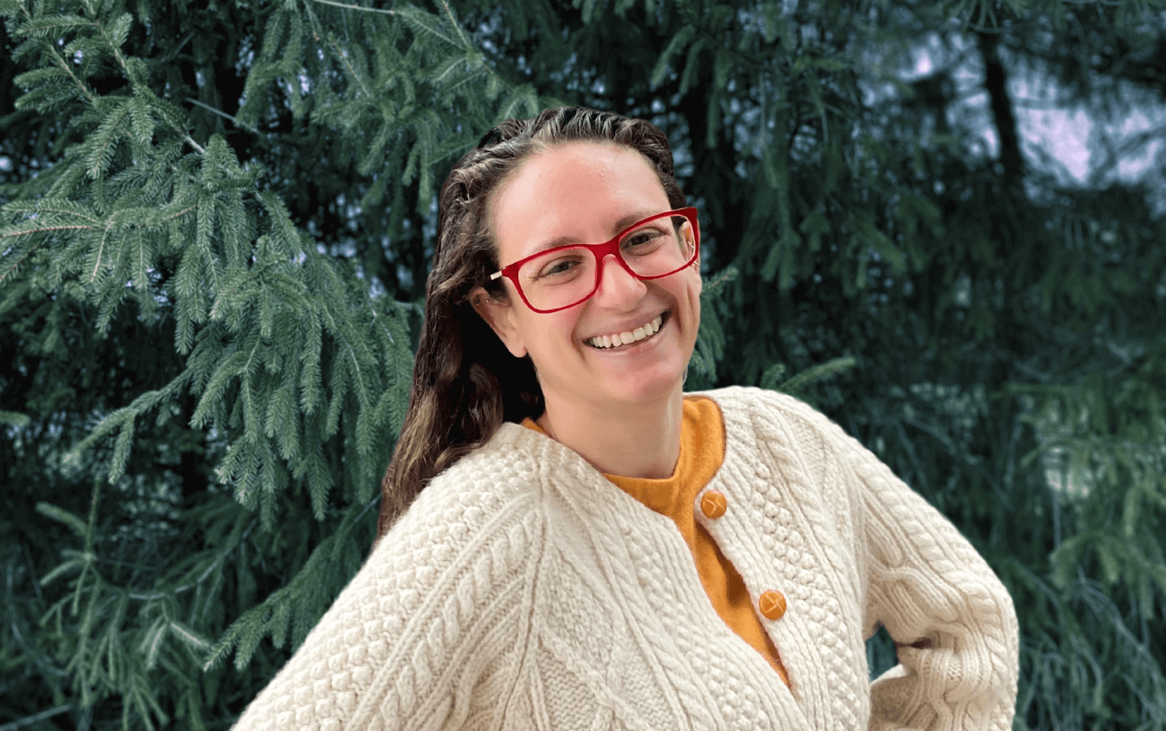 A smiling woman in red glasses and a cream cable-knit sweater stands in front of green pine trees.