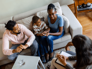 A family consults with a family therapist, sitting on a couch, while the mother holds a tablet and a child looks on. Consulting a family therapist about how family therapy can be helpful starts by finding a family therapist near you.