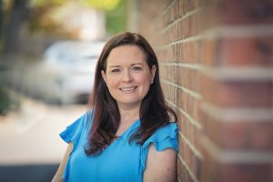 A woman with long dark hair in a blue shirt is smiling while leaning against a brick wall outside.