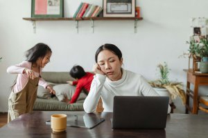 A tired mother with her head resting on her hand sits at a desk with a laptop while two children play on a couch in the background. The image captures the reality of balancing work and family life, especially relevant for neurodivergent moms seeking support through specialized counseling services in Branchburg, NJ.