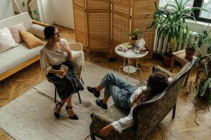 Two individuals in a counseling session in Branchburg NJ are seated in comfortable chairs having a conversation. The cozy space features natural light, a wooden divider, and multiple houseplants creating a calming environment for motherhood support and neurodivergent-affirming therapy Near Somerville NJ
