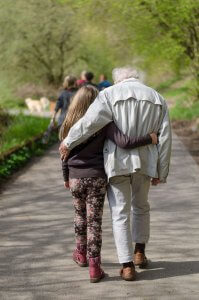 A young girl and her elderly grandfather walking arm-in-arm along a nature path surrounded by greenery. This represents the important role grandparents play in understanding and advocating for food allergy anxiety in Branchburg NJ. Therapy and counseling services help educate elders about safely managing their grandchildren's food allergies during family outings