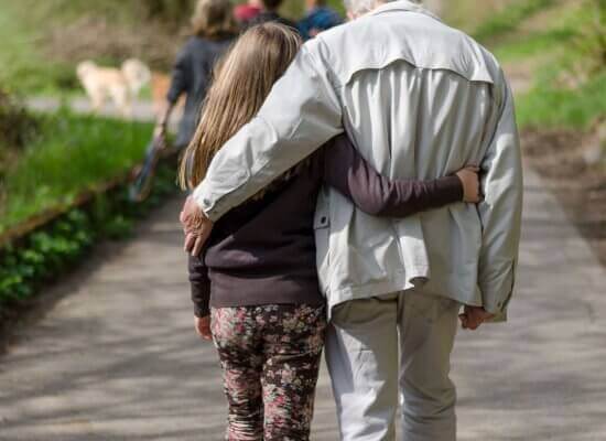 A young girl and her elderly grandfather walking arm-in-arm along a nature path surrounded by greenery. This represents the important role grandparents play in understanding and advocating for food allergy anxiety in Branchburg NJ. Therapy and counseling services help educate elders about safely managing their grandchildren's food allergies during family outings
