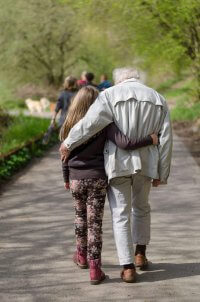 A young girl and her elderly grandfather walking arm-in-arm along a nature path surrounded by greenery. This represents the important role grandparents play in understanding and advocating for food allergy anxiety in Branchburg NJ. Therapy and counseling services help educate elders about safely managing their grandchildren's food allergies during family outings