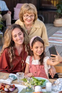 Three generations—grandmother, mother, and daughter—smiling together and taking a selfie at an outdoor dining table with fresh food, representing family unity in managing food allergies. Therapy services in Branchburg NJ provide resources for educating grandparents about recognizing food allergy symptoms and creating safe dining experiences for multi-generational gatherings
