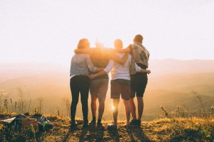 backlit photograph showing a group of four people standing with arms around each other on a hilltop at sunset, facing away from the camera toward a golden horizon. The warm light creates silhouettes of their bodies as they share a moment of connection and solidarity, representing the healing power of community support for neurodivergent individuals in Scotch Plains NJ